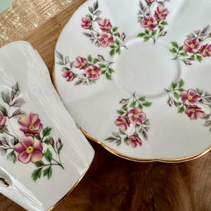 May include: A white teacup and saucer set with a delicate floral pattern. The set features pink and green flowers on a white background with a gold trim.