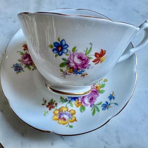 Puede incluir: Juego de taza y platillo blancos con un dise&ntilde;o floral con rosas rosas, flores azules y amarillas y hojas verdes. Los bordes de la taza y el platillo son festoneados y ribeteados con oro.