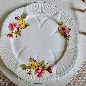 May include: White ceramic plate with a scalloped edge and a light blue rim. The plate is decorated with floral clusters of pink and yellow flowers with green leaves. The plate is on a heart-shaped tray.