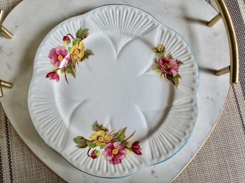 May include: White ceramic plate with a scalloped edge and a light blue rim. The plate is decorated with floral clusters of pink and yellow flowers with green leaves. The plate is on a heart-shaped tray.