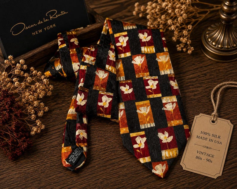Op de afbeelding: Een vintage zijden stropdas van Oscar de la Renta, met een herhalend bloemmotief in rood, oranje en wit op een zwarte achtergrond. Het label luidt "100% Zijde Made in USA Vintage 80s-90s".