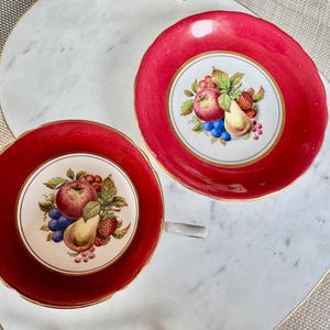 May include: Two red teacups with gold trim and a white floral design featuring fruit, including apples, pears, and grapes.