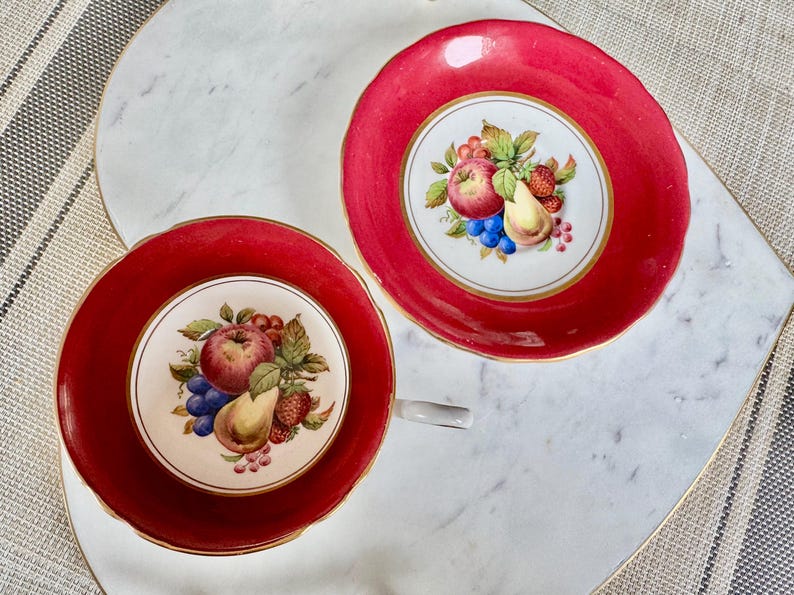 May include: Two red teacups with gold trim and a white floral design featuring fruit, including apples, pears, and grapes.