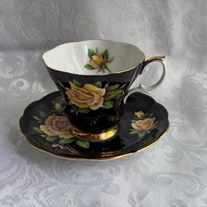 Pode incluir: Um conjunto de ch&aacute;vena e pires em porcelana preta com um padr&atilde;o de rosas amarelas vibrantes e folhas verdes. A ch&aacute;vena tem um rebordo recortado, uma asa com acabamento dourado e uma base dourada. O interior exibe um bot&atilde;o de rosa amarelo.