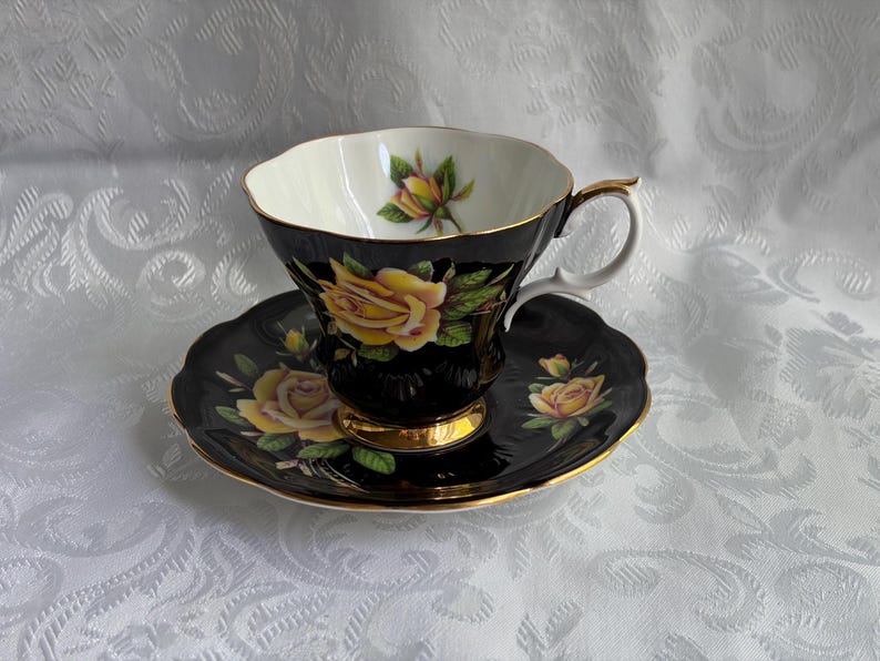 Pode incluir: Um conjunto de ch&aacute;vena e pires em porcelana preta com um padr&atilde;o de rosas amarelas vibrantes e folhas verdes. A ch&aacute;vena tem um rebordo recortado, uma asa com acabamento dourado e uma base dourada. O interior exibe um bot&atilde;o de rosa amarelo.