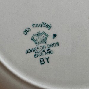 May include: Close-up of a white ceramic plate with a teal-colored emblem. The emblem features a crown and the words "Old English" and "Johnson Bros Made in England BY". The plate has a slightly curved edge.