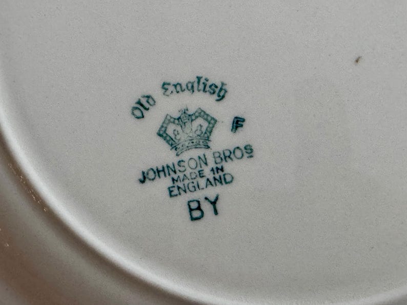 May include: Close-up of a white ceramic plate with a teal-colored emblem. The emblem features a crown and the words "Old English" and "Johnson Bros Made in England BY". The plate has a slightly curved edge.