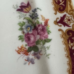 May include: A white ceramic plate with a floral design. The design features pink roses, purple and yellow flowers, and green leaves. The plate has a gold and burgundy border.