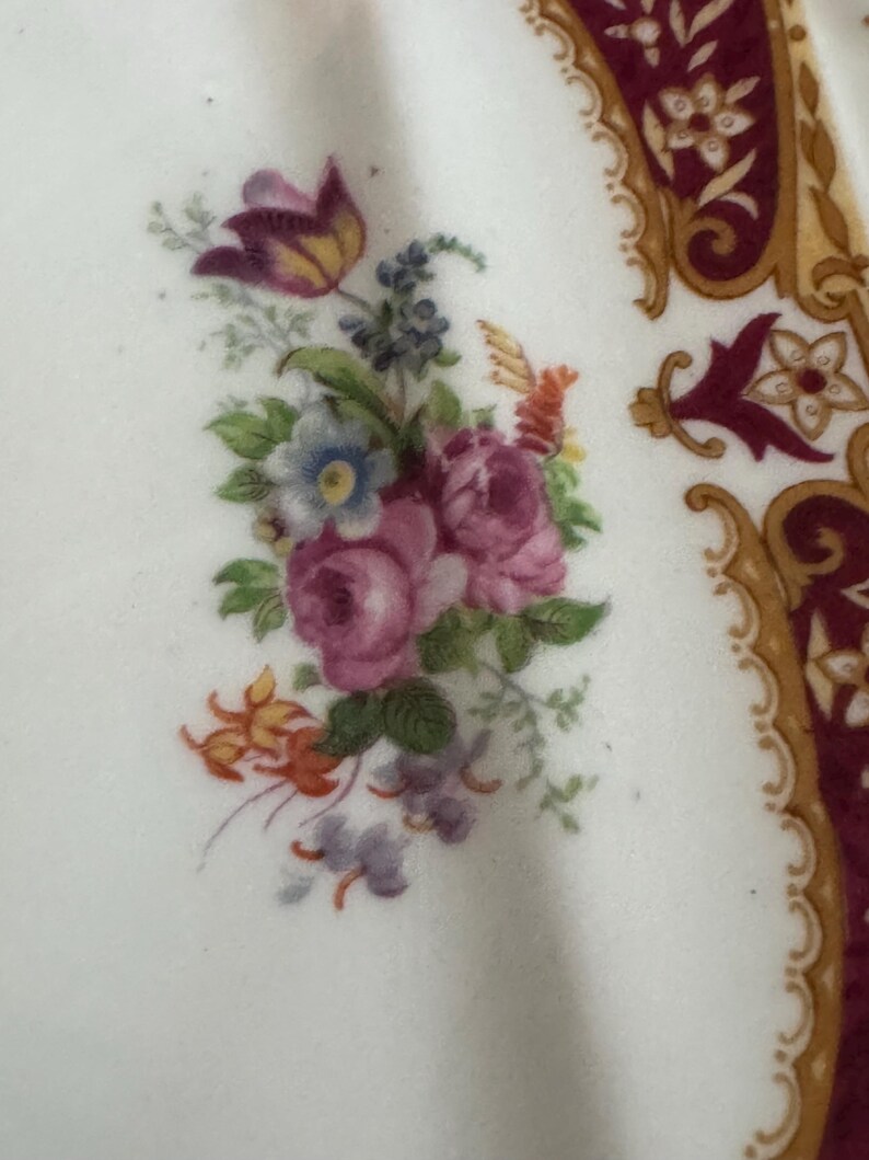 May include: A white ceramic plate with a floral design. The design features pink roses, purple and yellow flowers, and green leaves. The plate has a gold and burgundy border.