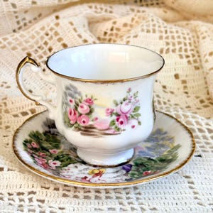May include: A white porcelain teacup and saucer set featuring a delicate floral pattern with pink roses and green foliage. The cup has a scalloped rim and a gold-trimmed handle, while the saucer displays a matching garden scene with gold edges.