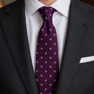 May include: A dark purple necktie featuring a repeating pattern of small white paisley motifs, worn with a crisp white collared shirt and a charcoal gray suit jacket that includes a white pocket square tucked into the breast pocket.