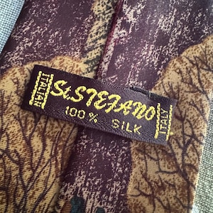 Pu&ograve; includere: Una cravatta in 100% seta con un motivo astratto bordeaux scuro e marrone con sagome di alberi. Un'etichetta rettangolare in tessuto &egrave; cucita sul retro, riportante il testo St. Stefano, 100% Silk e Italian.