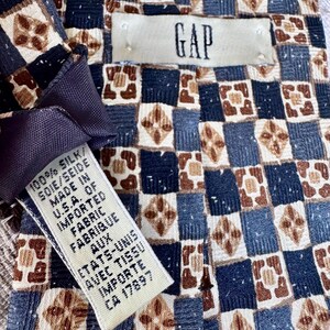 May include: A necktie featuring a repeating geometric checkerboard pattern in shades of navy blue, light blue, and tan. The tie includes a GAP brand label and a care tag stating 100% silk, made in U.S.A. of imported fabric.