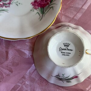 May include: A white bone china teacup and saucer set featuring pink carnation floral patterns and gold trim. The underside of the teacup base displays the text Queen Anne BONE CHINA ENGLAND with a crown icon above the brand name.