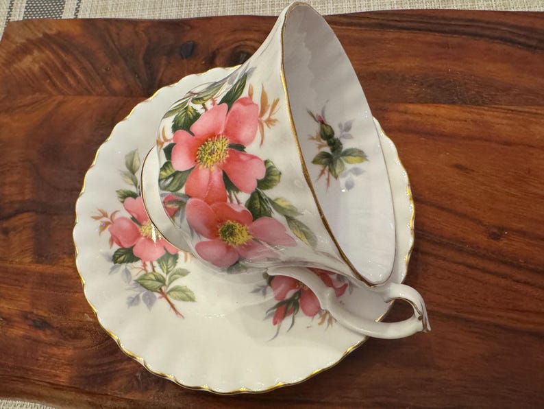 Puede incluir: Una taza de t&eacute; y un platillo blancos con un dise&ntilde;o floral con rosas rosas y hojas verdes. La taza y el platillo tienen un borde dorado.
