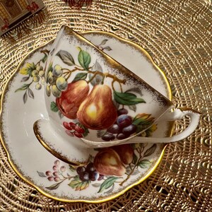 Op de afbeelding: Een wit theekopje en schotel met een gouden rand. Het kopje en de schotel zijn versierd met een bloemmotief met peren, druiven en ander fruit. De achtergrond is een gouden kantkleed.