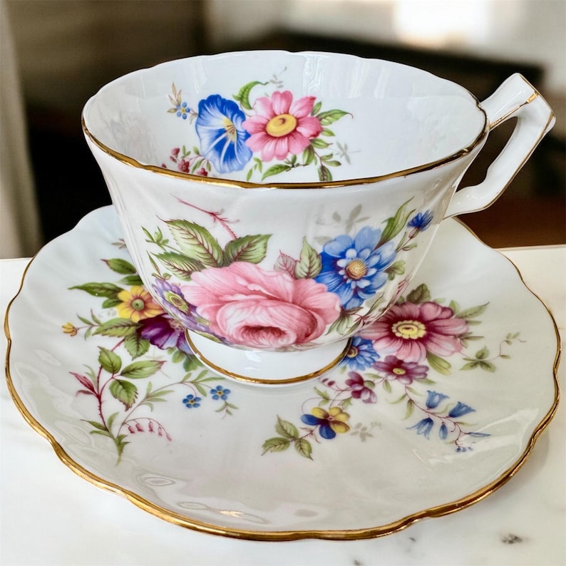May include: A white teacup and saucer with a floral pattern. The cup and saucer have a gold trim. The flowers are pink, blue, and yellow.
