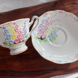 May include: A white teacup and saucer set with a floral design. The cup and saucer feature a colorful floral pattern with pink, yellow, blue, and green flowers.