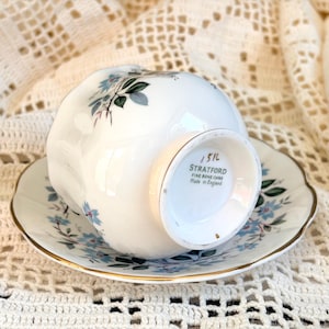 May include: A white fine bone china teacup and saucer set featuring a delicate blue floral pattern with green leaves and gold trim. The base of the cup is marked with the text Stratford Fine Bone China Made in England and the number 1516.