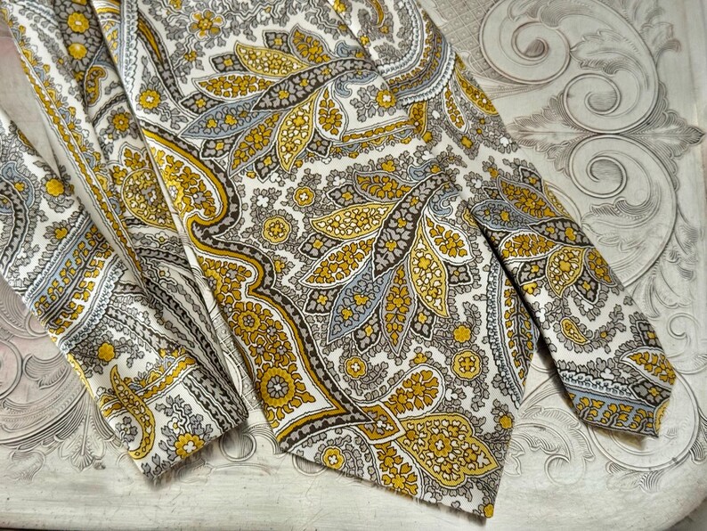 May include: A vintage paisley tie and matching pocket square. The tie features a repeating pattern of yellow, gray, and brown paisley shapes on a white background. The pocket square is folded and visible next to the tie.