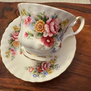 May include: A white teacup and saucer with a floral pattern. The cup and saucer have a gold trim. The flowers are pink, red, yellow, and blue.