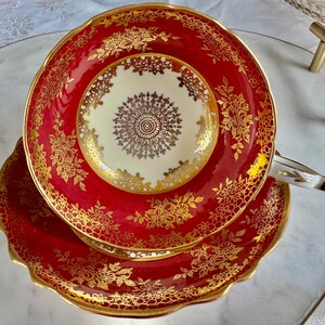 May include: A red and gold teacup and saucer set. The teacup and saucer have a scalloped edge and are decorated with gold floral patterns. The center of the teacup is cream-colored with a gold design. The handle is white with gold accents.