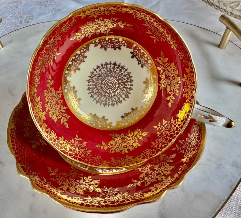 May include: A red and gold teacup and saucer set. The teacup and saucer have a scalloped edge and are decorated with gold floral patterns. The center of the teacup is cream-colored with a gold design. The handle is white with gold accents.