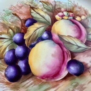 May include: A decorative plate featuring a still life of fruit. The image showcases ripe purple grapes, peaches with pink and yellow hues, and green leaves. The plate has a cream-colored border with a detailed design.