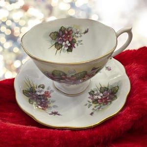 May include: A white teacup and saucer set with a floral design featuring purple and pink flowers. The set has a gold trim around the edges.