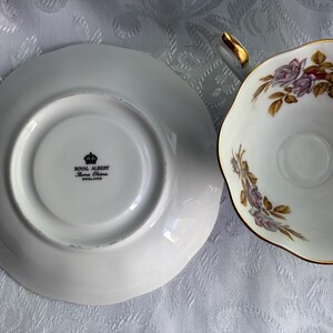 Może przedstawiać: Zestaw filiżanka ze spodkiem z porcelany kostnej Royal Albert. Na spodku widnieje czarny znak firmowy Royal Albert Bone China England. Filiżanka ozdobiona jest delikatnym motywem kwiatowym z fioletowymi r&oacute;żami, zielonymi liśćmi i złotym brzegiem.