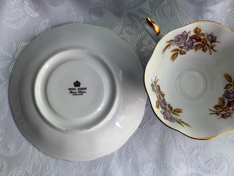 Może przedstawiać: Zestaw filiżanka ze spodkiem z porcelany kostnej Royal Albert. Na spodku widnieje czarny znak firmowy Royal Albert Bone China England. Filiżanka ozdobiona jest delikatnym motywem kwiatowym z fioletowymi r&oacute;żami, zielonymi liśćmi i złotym brzegiem.