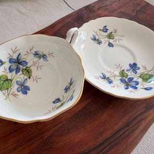 May include: A white teacup and saucer set with a delicate floral pattern featuring blue and green violets. The set has a gold trim around the edges.
