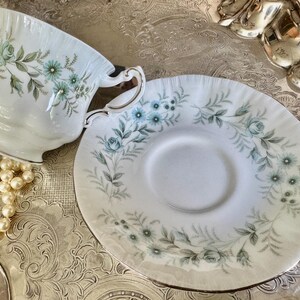 May include: A white teacup and saucer set with a floral design in shades of blue and green. The teacup has a gold rim and handle. The saucer is round with a matching floral pattern. The set is on a silver tray, with a pearl necklace.