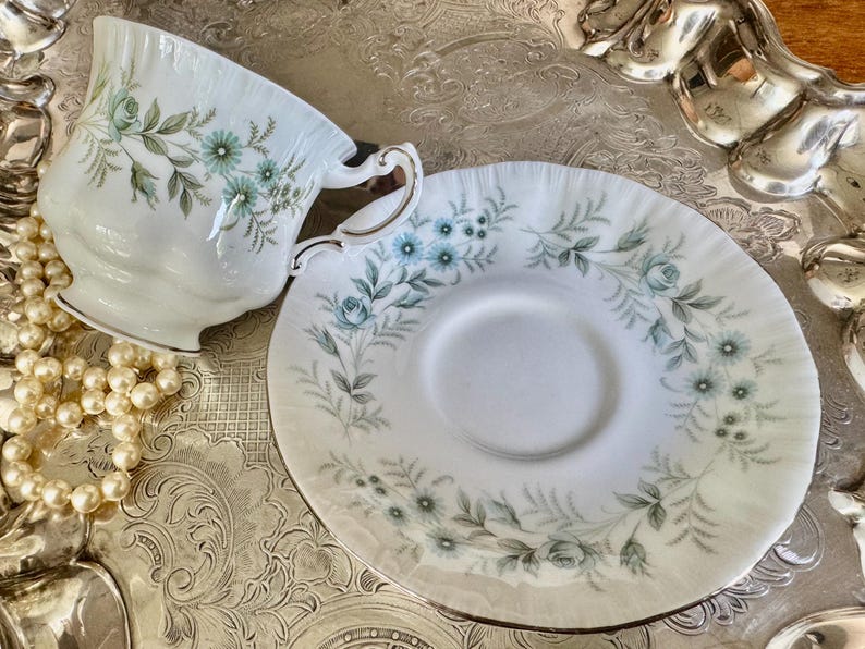 May include: A white teacup and saucer set with a floral design in shades of blue and green. The teacup has a gold rim and handle. The saucer is round with a matching floral pattern. The set is on a silver tray, with a pearl necklace.