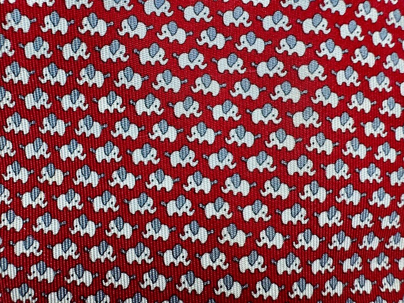 Puede incluir: Una corbata roja con un patr&oacute;n repetitivo de peque&ntilde;os elefantes blancos con orejas de color azul claro. La tela tiene una textura acanalada diagonal visible en toda la superficie, creando un dise&ntilde;o consistente y uniforme.