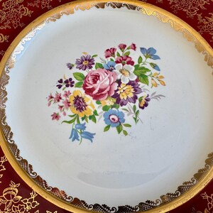 May include: A white ceramic plate with a gold rim and a floral bouquet design in the center. The bouquet features pink, purple, yellow, and blue flowers. The plate sits on a red tray with a gold floral pattern.