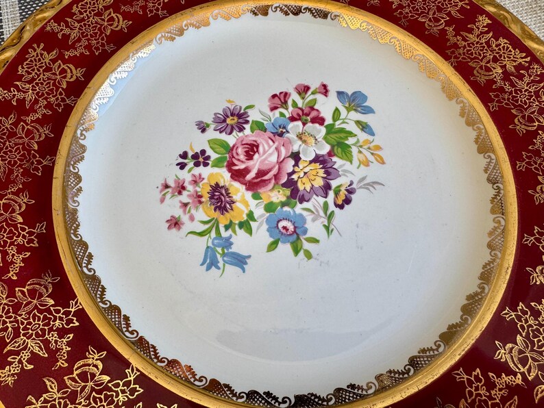 May include: A white ceramic plate with a gold rim and a floral bouquet design in the center. The bouquet features pink, purple, yellow, and blue flowers. The plate sits on a red tray with a gold floral pattern.