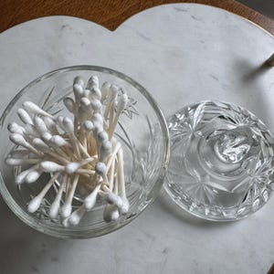 May include: A clear glass jar with a faceted starburst pattern containing a cluster of white cotton swabs. The jar has a matching removable lid with a central knob handle, showcasing a reflective, transparent design for storage.