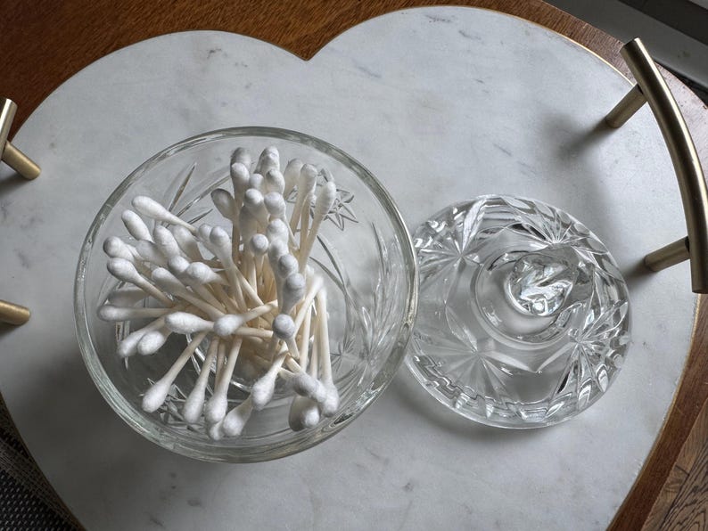 May include: A clear glass jar with a faceted starburst pattern containing a cluster of white cotton swabs. The jar has a matching removable lid with a central knob handle, showcasing a reflective, transparent design for storage.