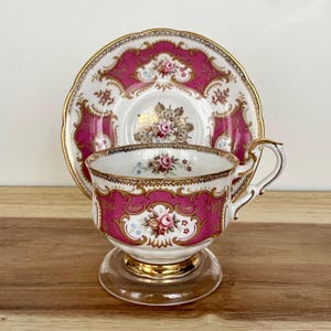 May include: A vintage teacup and saucer set. The set features a vibrant pink and gold design with floral accents. The teacup has a clear glass base and a gold rim. The saucer mirrors the cup's design, creating a cohesive and elegant look.