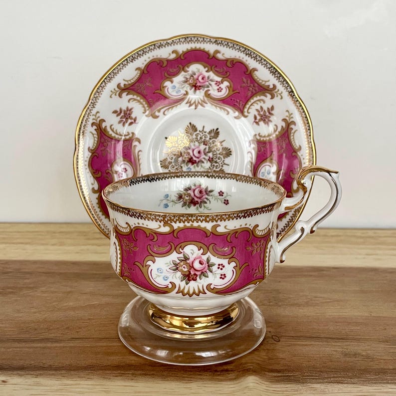 May include: A vintage teacup and saucer set. The set features a vibrant pink and gold design with floral accents. The teacup has a clear glass base and a gold rim. The saucer mirrors the cup's design, creating a cohesive and elegant look.