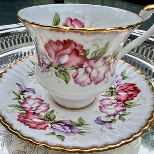 May include: White teacup and saucer set with a floral design in shades of pink, red, and purple. The edges are trimmed with gold. The teacup is sitting on a silver tray.