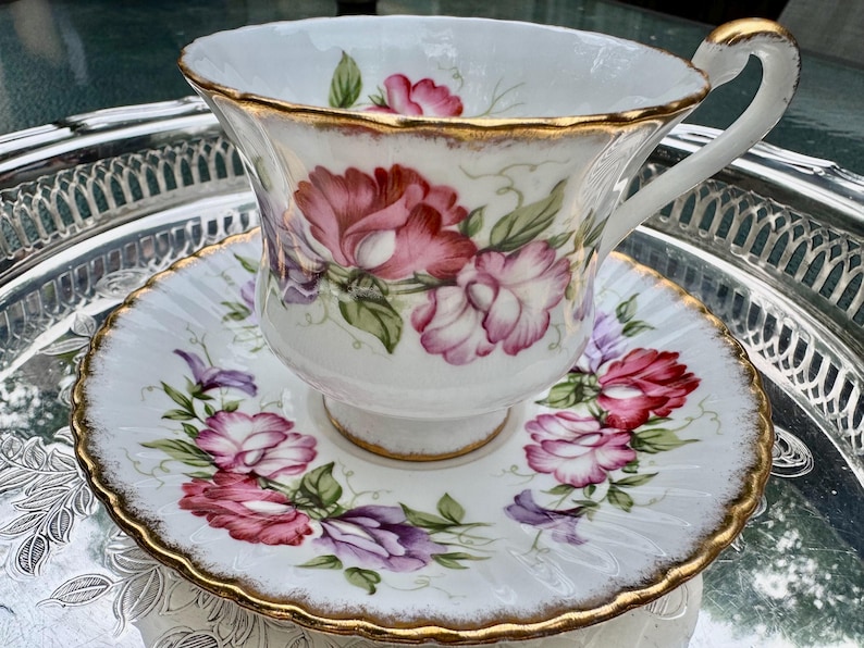 May include: White teacup and saucer set with a floral design in shades of pink, red, and purple. The edges are trimmed with gold. The teacup is sitting on a silver tray.