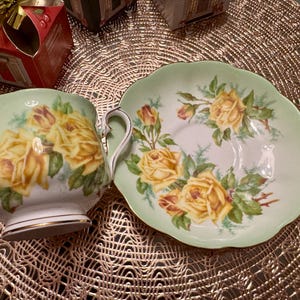May include: A green and white teacup and saucer set with a floral design featuring yellow roses. The teacup has a handle and the saucer has a scalloped edge.