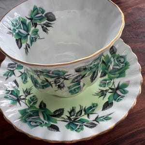 May include: A white teacup and saucer with a green and white floral pattern. The cup has a gold rim and the saucer has a scalloped edge.