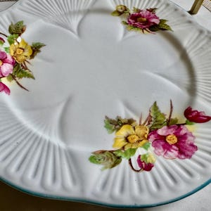 May include: White ceramic serving plate with a scalloped edge and a floral design. The plate features pink and yellow flowers with green leaves. The rim has a light blue trim and a ribbed texture.