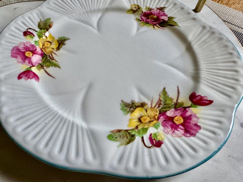 May include: White ceramic serving plate with a scalloped edge and a floral design. The plate features pink and yellow flowers with green leaves. The rim has a light blue trim and a ribbed texture.