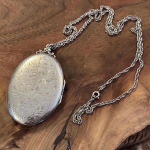May include: Silver oval locket necklace with an ornate floral design. The locket hangs from a delicate chain with a lobster clasp. The locket is approximately 1.5 inches long and 1 inch wide.
