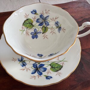 May include: A white teacup and saucer with a delicate floral pattern featuring blue and green violets. The cup has a gold rim and a handle. The saucer has a matching floral design.