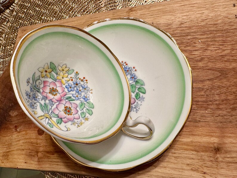 May include: A vintage teacup and saucer set with a green and white floral pattern. The cup and saucer have a gold trim around the edges.